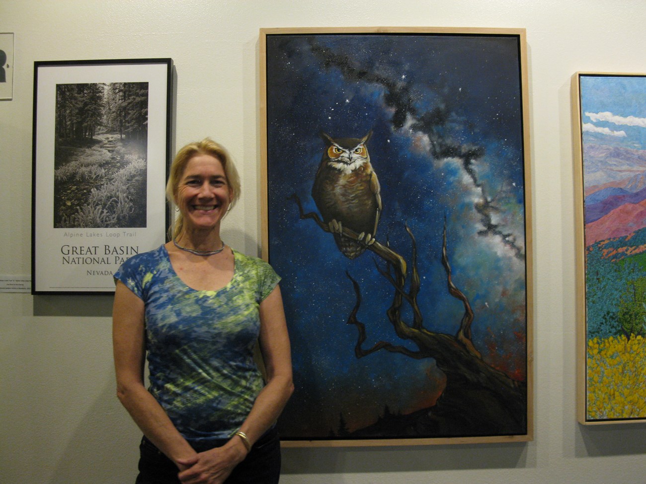 A smiling woman stands next to a painting of an owl sitting on a tree branch with the milky way behind it. The woman stands with hands clasped in front of her, smiling at the camera with pride.