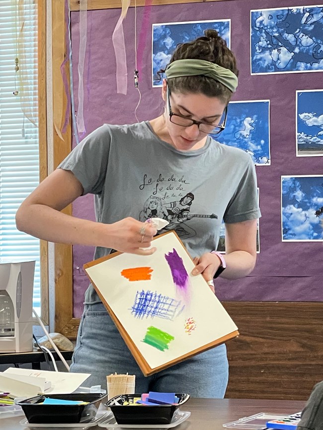 A woman stands holding a piece of paper with several paint colors on it. Paints have been applied different ways to the paper, and the woman looks down and points intentionally at one patch of color.