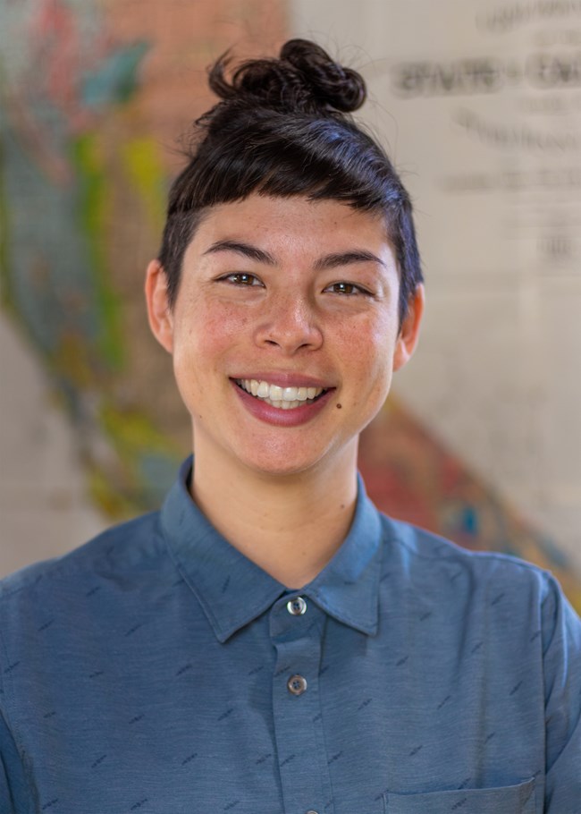 A person with dark hair in a high bun with very short bangs and shaved sides of their head. They are wearing a collared blue shirt, smiling in front of a geologic map of California.