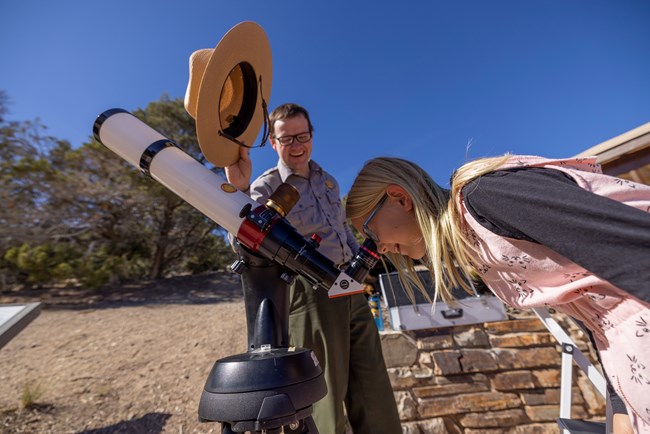 A ranger holds a hat to block the sun from a young girl looking through a solar telescope