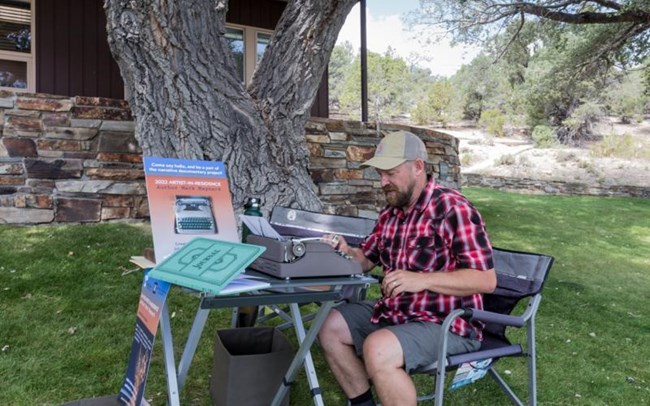 A man sits in a camp chair on a lawn with a typewriter on a folding table in front of him. An open book labeled journal sits behind the rypewriter with a sign stating "2022 Artist-in-Residence"