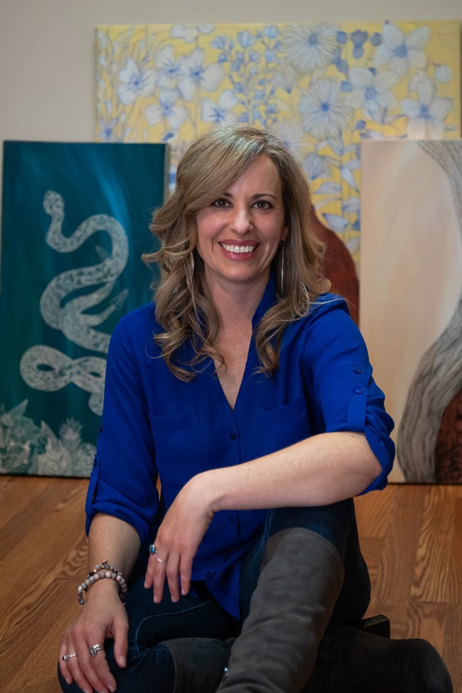 A color photo portrait of a smiling woman looking directly at the camera. Her dirty blonde hair reaches below her shoulders, wears a blue v-neck shirt, and sits in front of several highly detailed art pieces behind her.