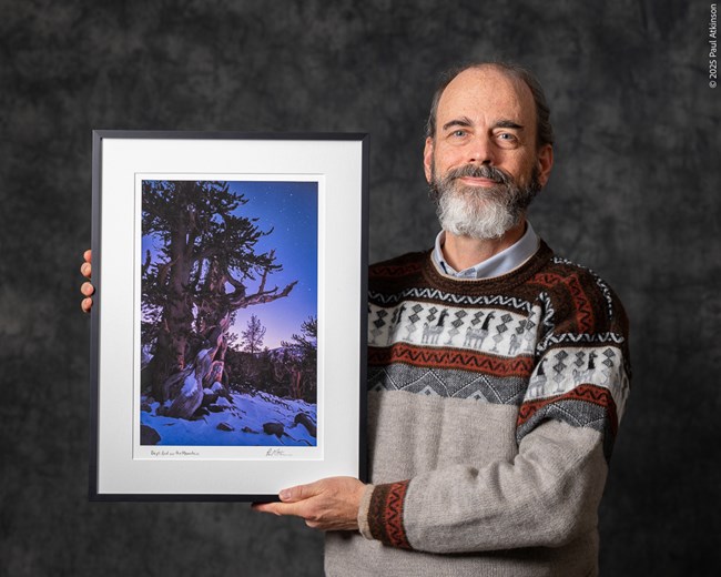 A smiling man stands holding a framed photograph. He stands straight, presenting the photo to the viewer, wearing a cozy sweater with alpacas and geometric patterns. The photo is of a gnarly tree in front od a dusky, star studded sky