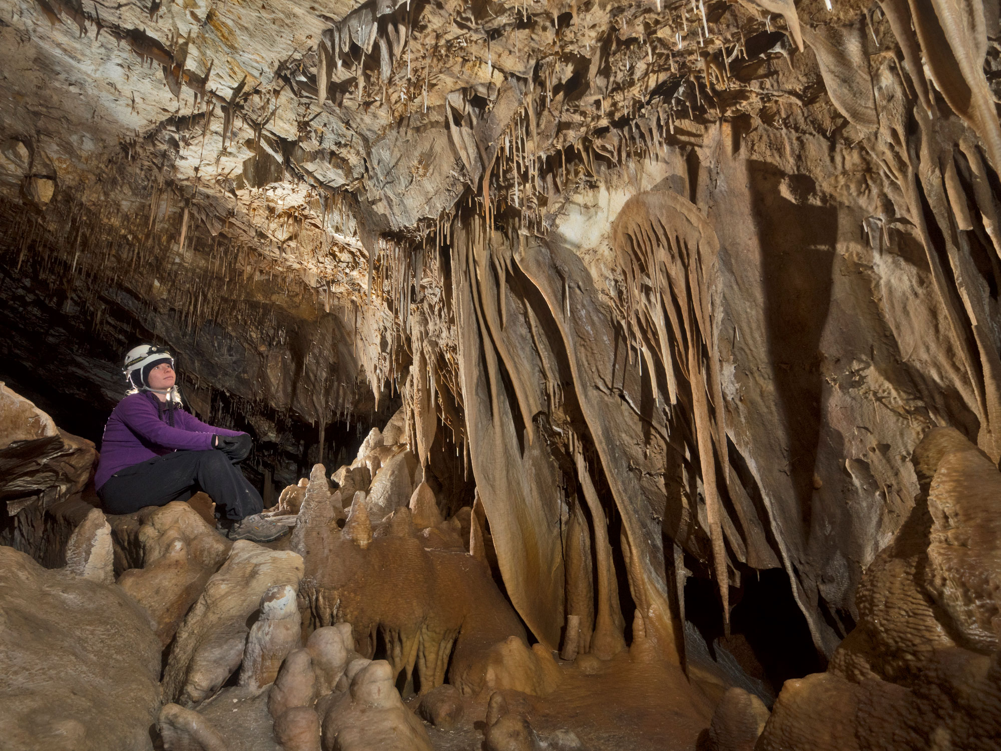 A caver sits on a rock shining her headlamp onto drapery formations cascading down a rock wall.