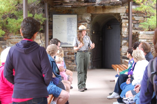A park ranger in uniform appears to be speaking to a group of visitors seated on benches outside of the entrance to a cave. The cave entrance has a closed door and a wall of stone surrounding it.