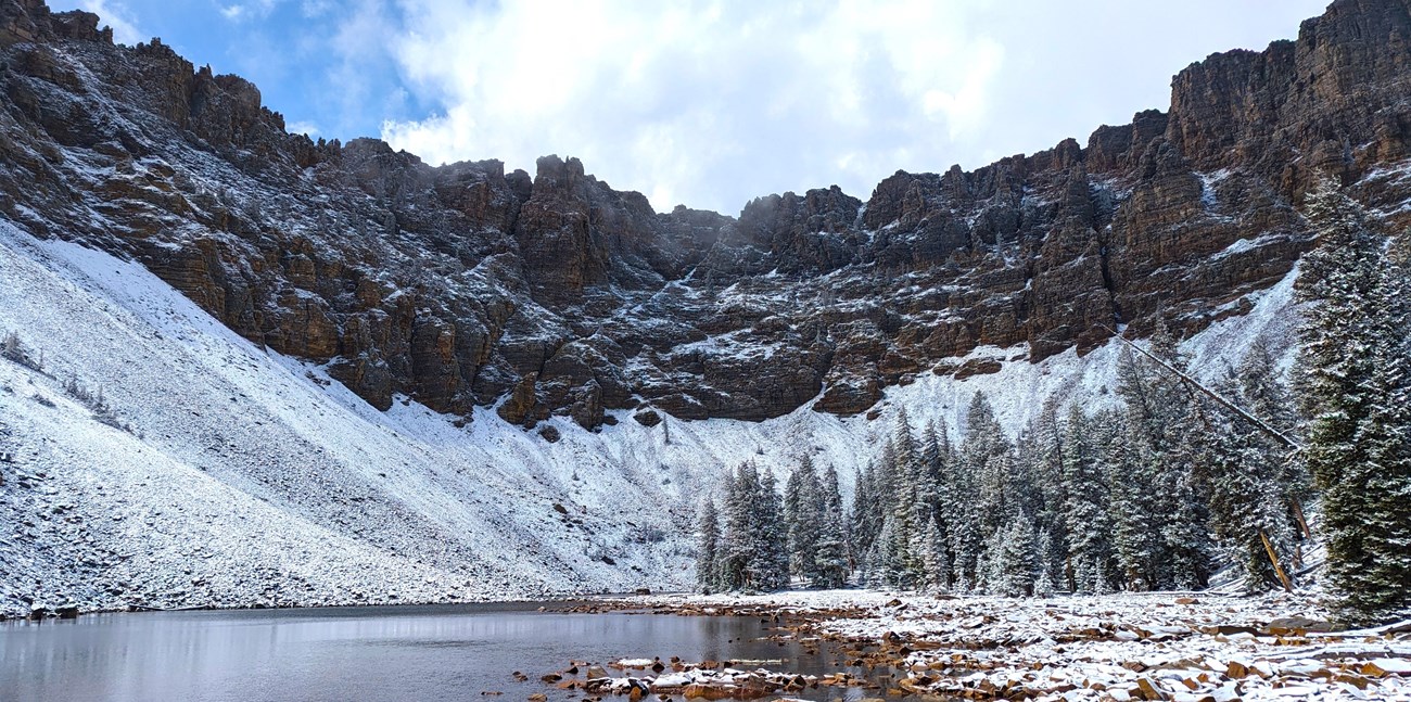 Jagged, rocky cliffs surround an alpine lake. snow covers the slopped bottoms of the cliffs, and a light layer surrounds the lake itself.