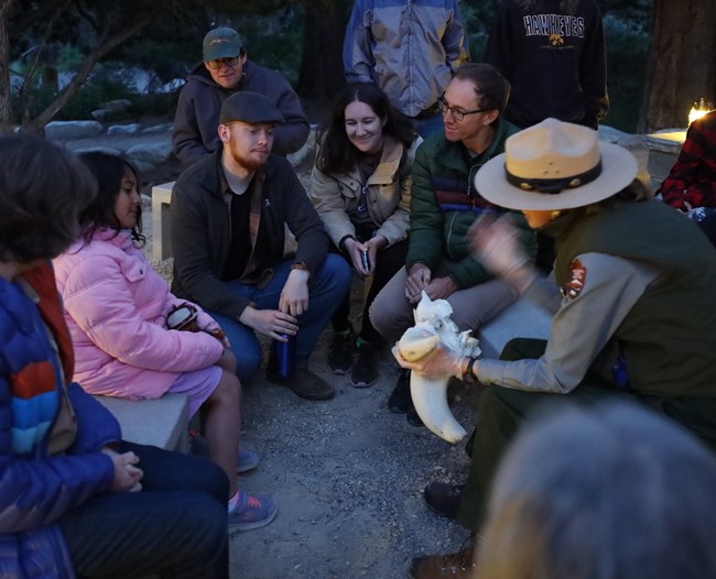 A color image of a group sitting in a cirlce, looking at a ranger with the slat brimmed straw hat holding a bighorn sheep skull