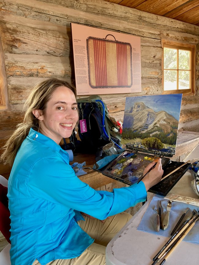 A smiling woman looks at the camera with a paintbursh in hand. A painting of mountains and trees is in front of her with an open palette beneath