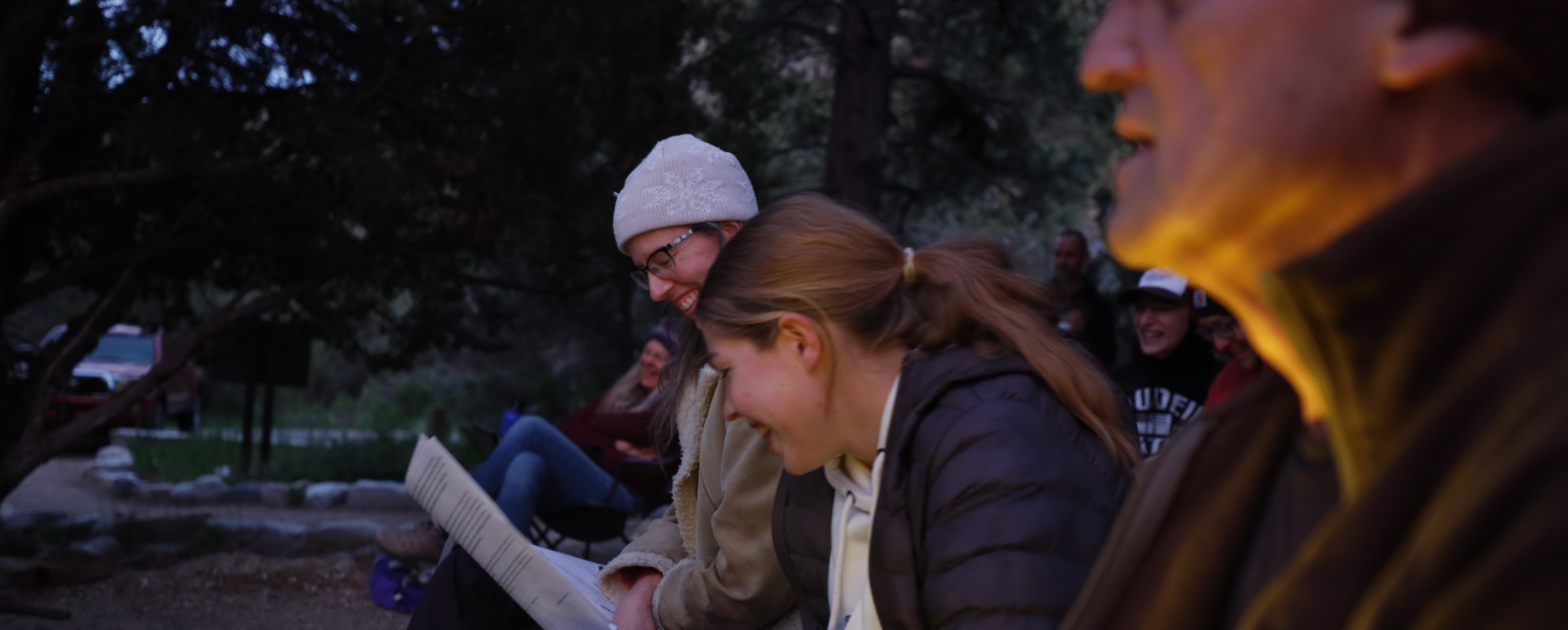 A color image with a thin line of focus on two smiling women looking down at a sheet of paper. Blurred faces in the background smile too in the glow of the evening.