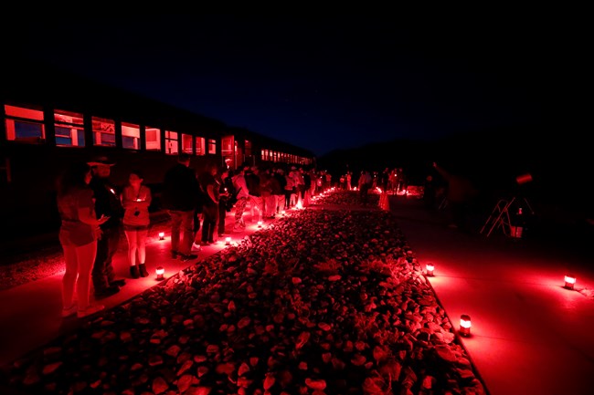 A crowded line stands around a concrete circle lit by red lanterns. Telescopes stand to one side, with a large train to the other, red light peering through the windows of the otherwise black shape. A small glow lights the sky with some small stars
