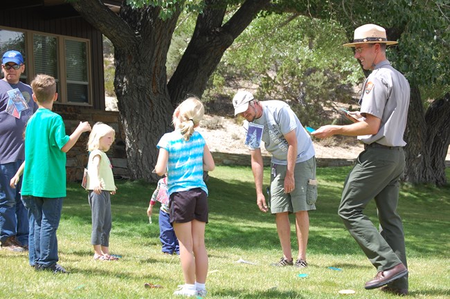 A park ranger stands to the right in a field of grass with kids and adults in a circle around him, and a large poplar tree in the background. The ranger directs kids and others in a playful activity, holding papers in various shapes and colors.