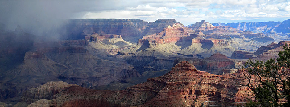 View east across the expanse of Grand Canyon in the morning. The right-hand side of the landscape is in sunlight. On the left, a thunderstorm with dark rain clouds is moving into the scene from the left. 