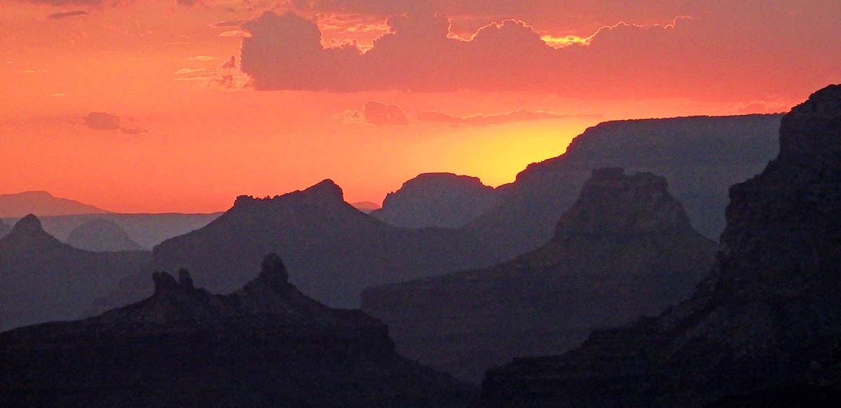 Looking west at sunset through a number of silhouettes of buttes and ridge lines within Grand Canyon. The sky, clouds, and everything in view is bathed in reddish-orange light. 