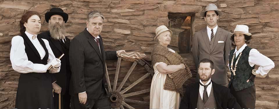 Wearing turn of the 19th Century clothing, 7 members of the Echoes From the Canyon living history production are posing in front of an adobe wall. A large wooden wagon wheel is in the center of the photo.