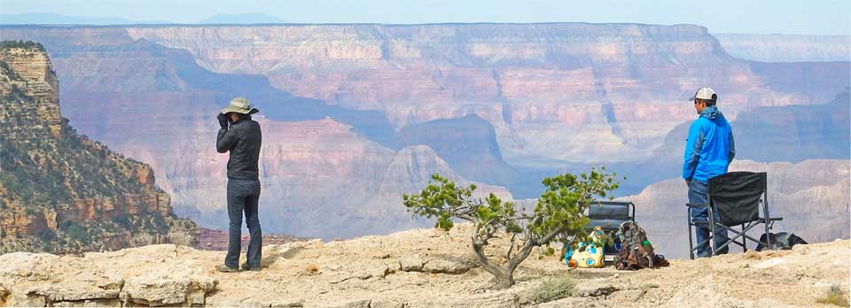 A man and a woman with binoculars standing on a rock ledge overlooking Grand Canyon. They are looking for raptors.