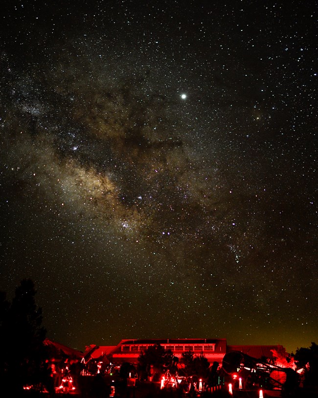 A starry night sky with part of the Milky Way visible above a telescope lot illuminated by red lights during the Grand Canyon National Park 2019 Star Party.