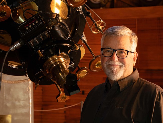 A man with glasses smiles in front of a telescope