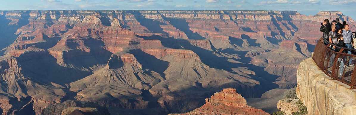 a group of sightseers behind a guardrail at a scenic overlook are viewing the vast and colorful Grand Canyon landscape.