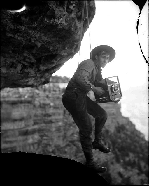 A man hangs down on a rope between two boulders holding a large, antique camera.
