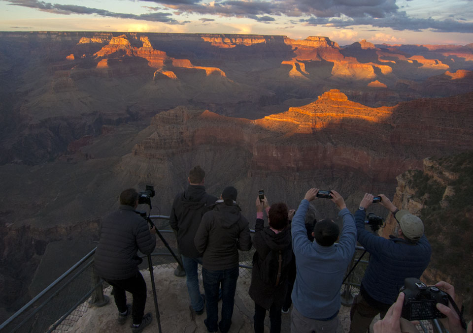 A group of visitors stand facing a sunset-lit canyon, photographing it with smart phones and traditional cameras.