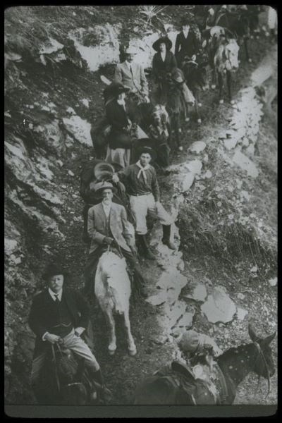 Looking up a trail, a black and white photo shows a group of travelers via mule on the trail at Grand Canyon.