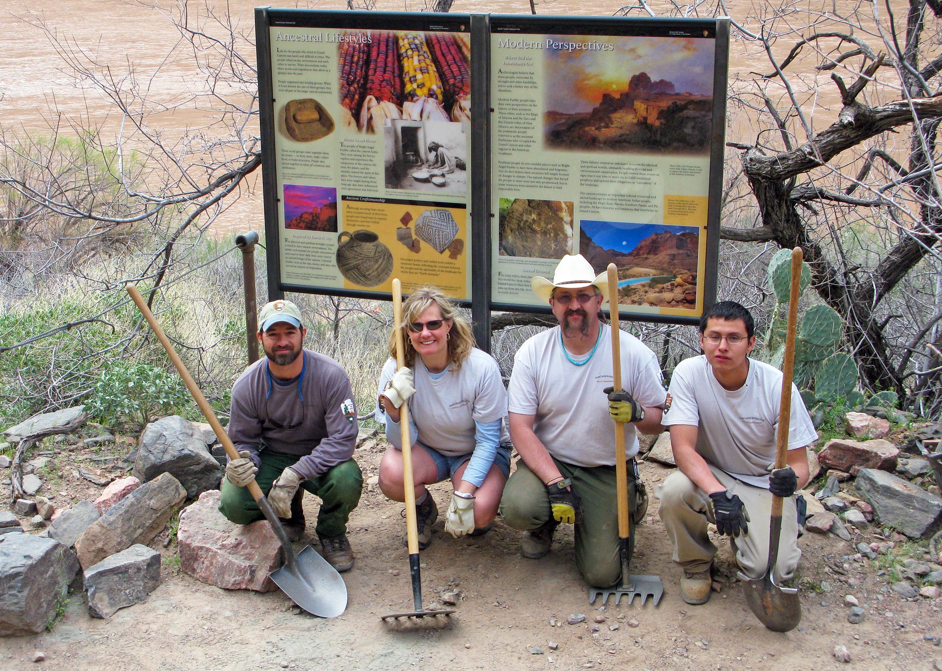 A group photo of 4 workers kneeling in front of two colorful outdoor exhibit panels interpreting an archaeology site. They are each posing while holding a shovel or a rake