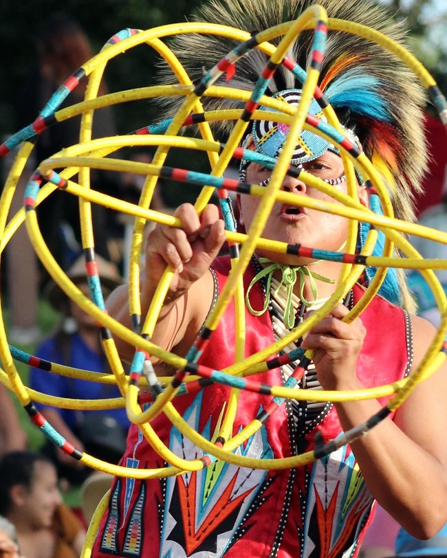 A man dressed in vibrant traditional regalia of blues, reds, yellows and white faces the camera holding yellow wrapped hoops as he dances.