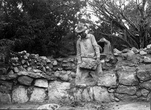 Black and white historic photo of a worker wearing coveralls placing a large block of stone onto the wall of a prehistoric structure.