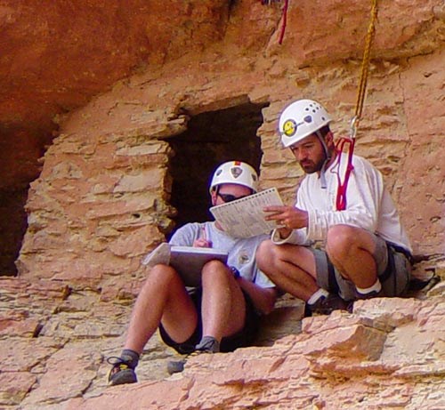 two archaeologists reviewing paperwork, are sitting in front of a stone granary with a door. They are wearing helmets and are roped into the cliff face.