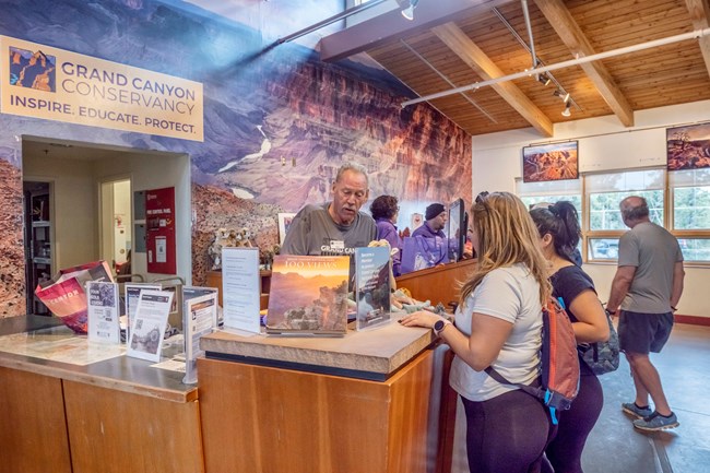 Several people stand at a cashier's desk, assisted by an employee; a sign above reads "Grand Canyon Conservancy."