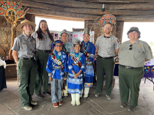 A picture of 4 members of Zuni Royalty in regalia standing together, 1 prince, 2 Princesses, and Miss Zuni with Park Rangers standing on either side of them. The photo was taken inside the base room of the Desert View Watchtower