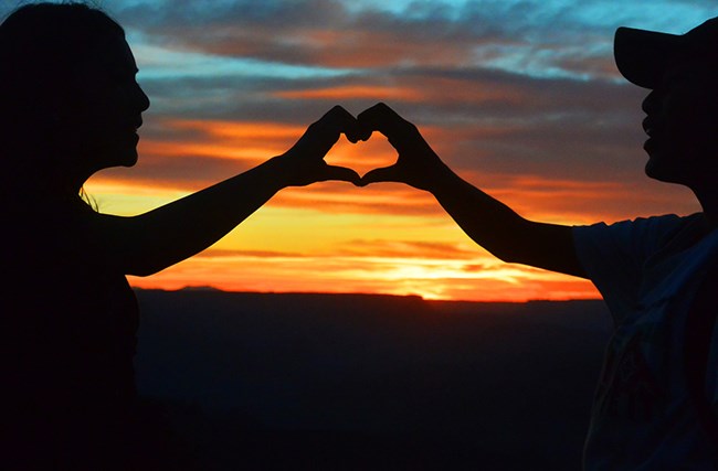 Two students are silhouetted by a multicolored sunset; students are making a heart shape with hands.
