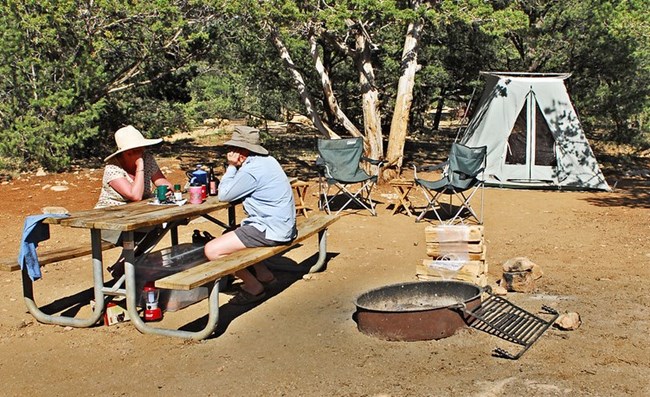 Two campers sit at a picnic table with an established fire ring close by.