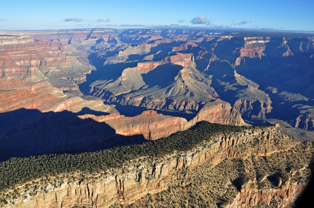 aerial view of a long, narrow plateau with sheer cliffs on all sides. In the background, colorful peaks and cliffs within a vast canyon landscape.