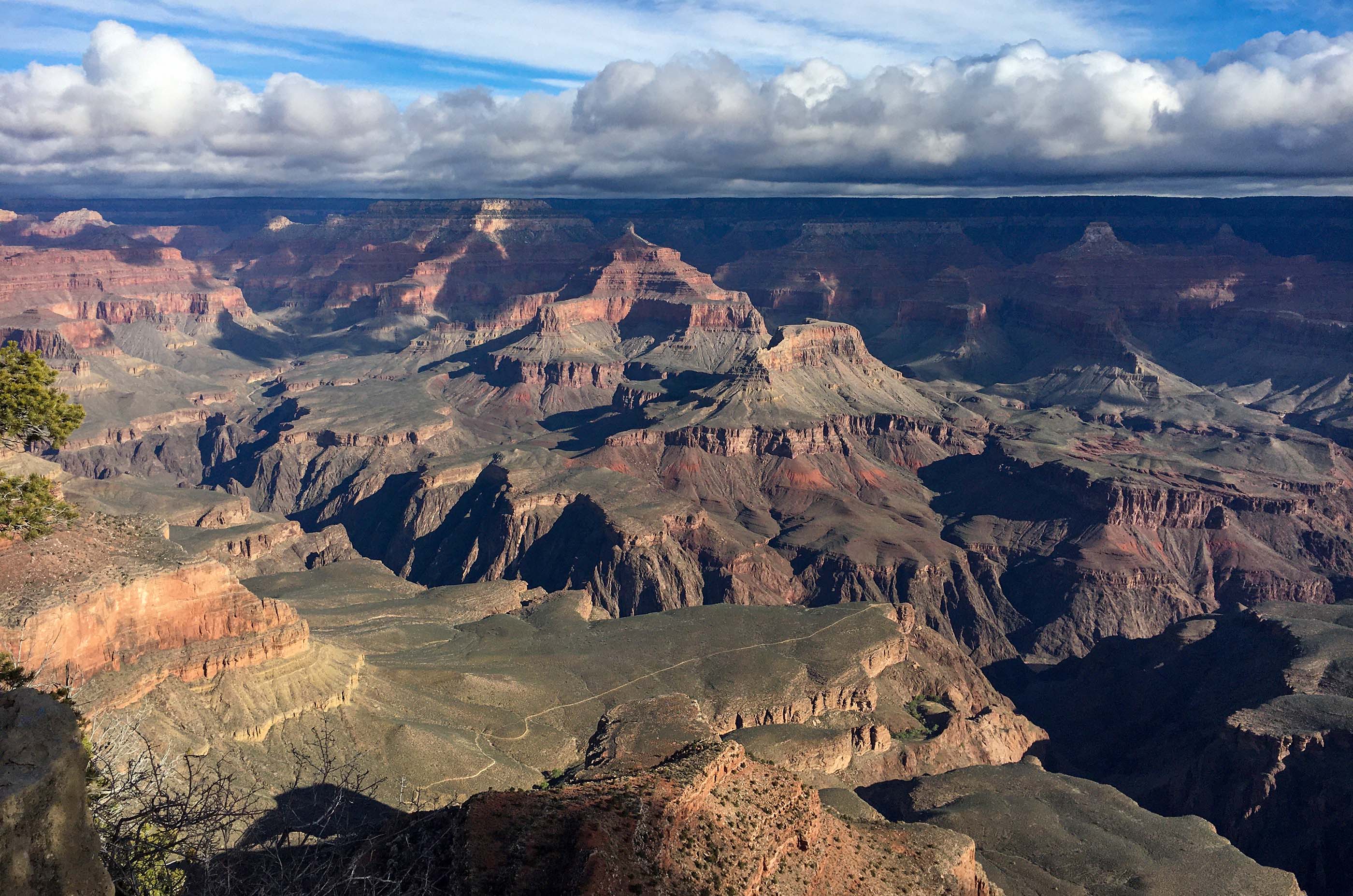 looking down across a plateau within a vast canyon with colorful peaks rising thousands of feet above. Threatening storm clouds shadow the distant rim.