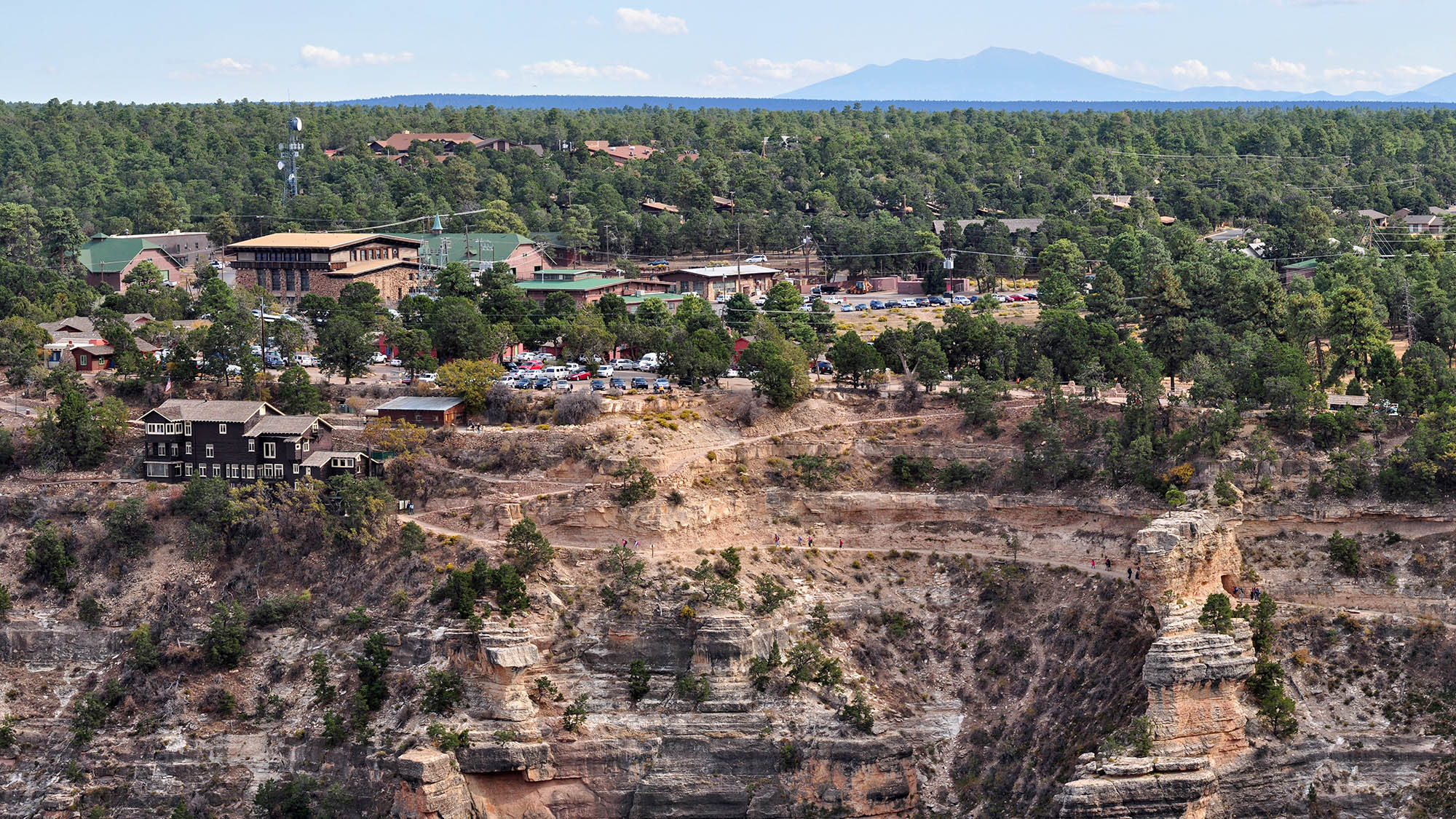 cars parked between buildings in a small village built on the edge of an almost sheer cliff. Several hundred feet of a backcountry trail is visible as it descends into the canyon.