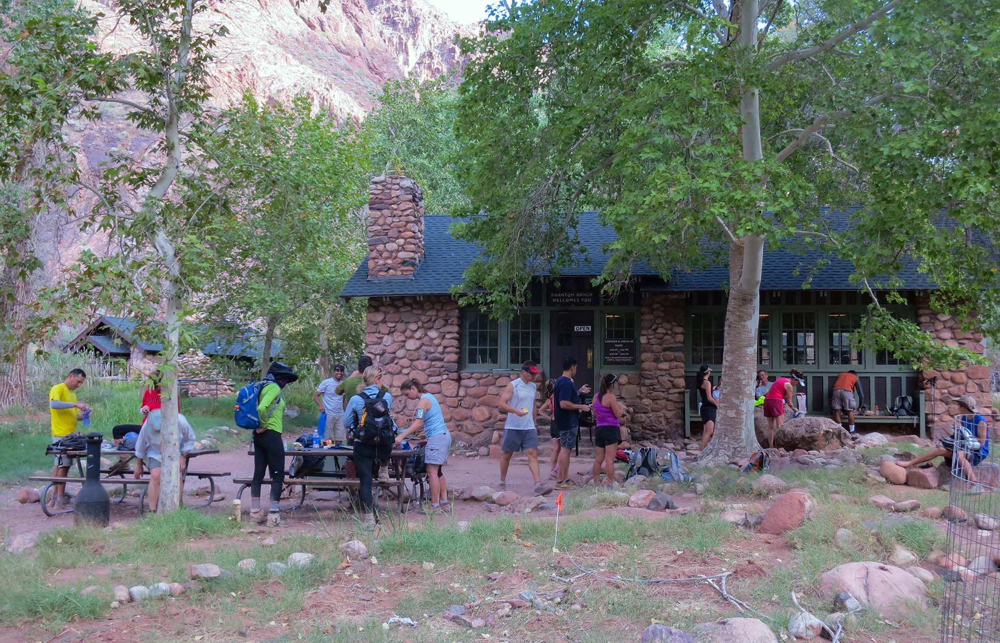 20 hikers and runners resting and organizing their gear in front of a rustic lodge built from river cobbles.