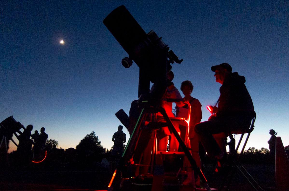 Twilight turning into night. An astronomer is letting people look through a large telescope, that is partially illuminated by red safety lights.