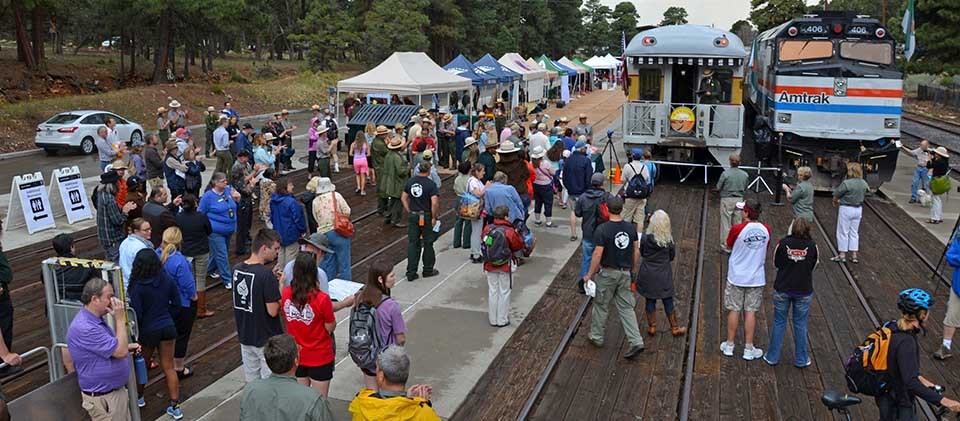 Colorful crowd of people at train depot listening to speeches. Two trains are on the right and to the left of trains is a row of exhibitor tents.