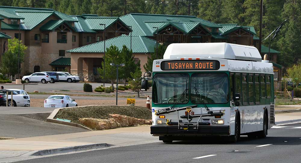 a white and green bus with a marque reading "Tusayan Route," driving past a large hotel several stories high with a green metal roof.