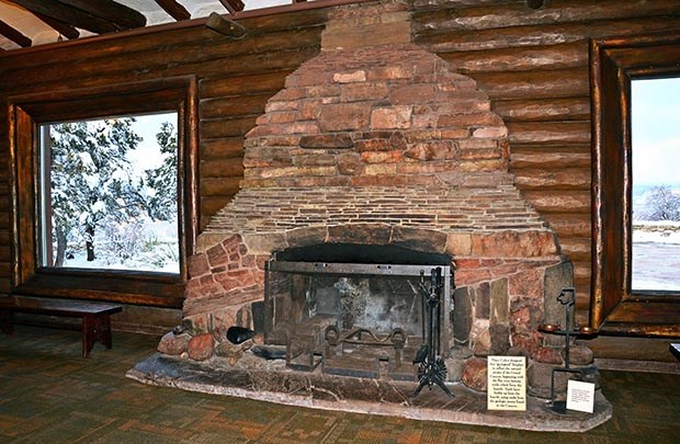 Against a log cabin style wall, a stone fireplace made from layers of rock strata is centered between two picture windows.
