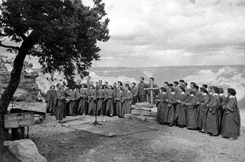 A choir wearing robes on either side of a stone altar and wooden cross. The Grand Canyon landscape is behind the choir.