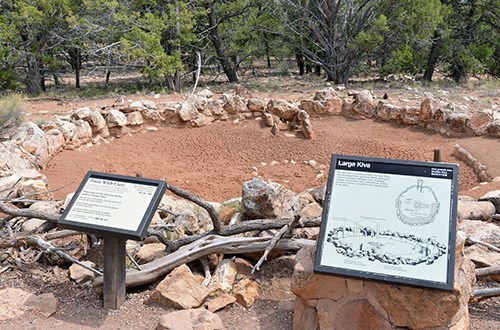 A stone wall circles around bare dirt with pine trees in the background, two informative signs in the foreground.