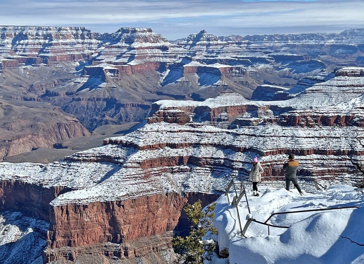 Two people standing behind metal guardrails at a snow covered scenic overlook; in the distance snow can be seen descending several thousand feet into a vast canyon covering stratified rock layers that form ridgelines and promontories.