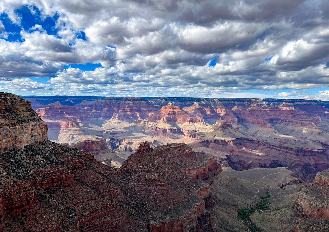 Almost filling the sky, a thick and clumpy layer of clouds is mostly shadowing colorful peaks and cliffs within a vast, mile deep canyon.