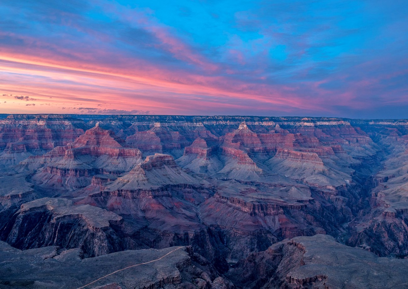 After sunset, as darkness is enveloping the landscape, brilliant pink clouds are reflecting pink light onto ridgelines, peaks and cliffs within a mile deep canyon.