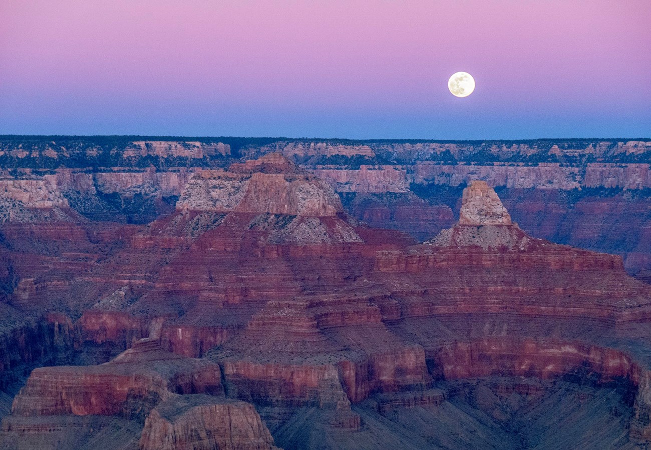 Just above the horizon, the full moon is rising in pink sky-glow above vermilion-colored peaks atop stratified cliffs within a vast canyon landscape.