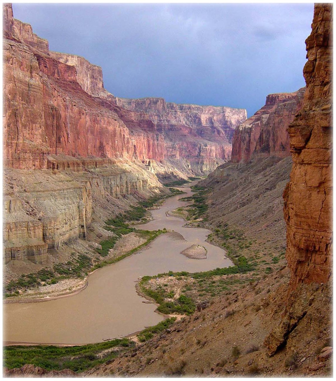Looking down from a canyon wall at a muddy Colorado River with colorful vermilion cliffs on both sides of the narrow gorge.