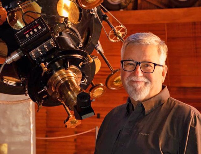 David Koerner with white hair and a neatly trimmed white beard and black gasses is standing below a large Refracting Telescope with numerous brass wheels and controls.