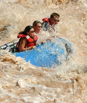 three individuals wearing orange life vests are riding in a blue raft through a muddy river rapid